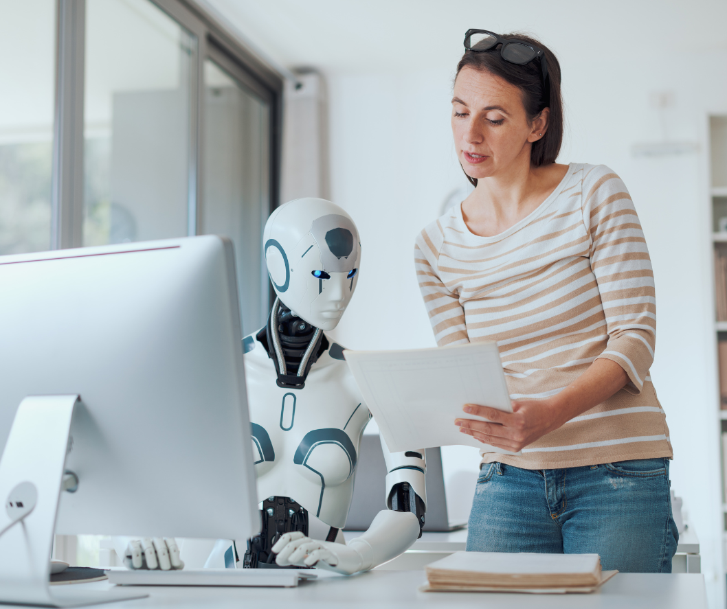 A white robot sits behind a computer, at a desk, while a human, female employee shows them something on a piece of paper.