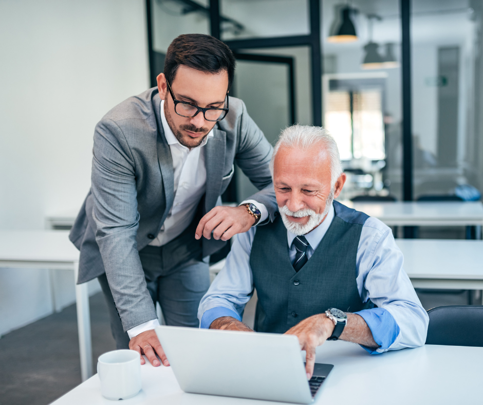 Younger executive leans over to discuss a salary‑sacrifice guide on a laptop with a senior colleague in a modern meeting room