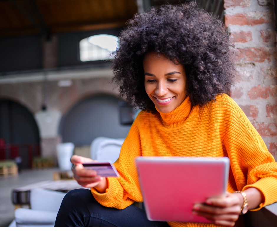 Smiling woman in a bright orange sweater holds a tablet and credit card in a loft‑style office, illustrating the seamless experience offered by tech schemes