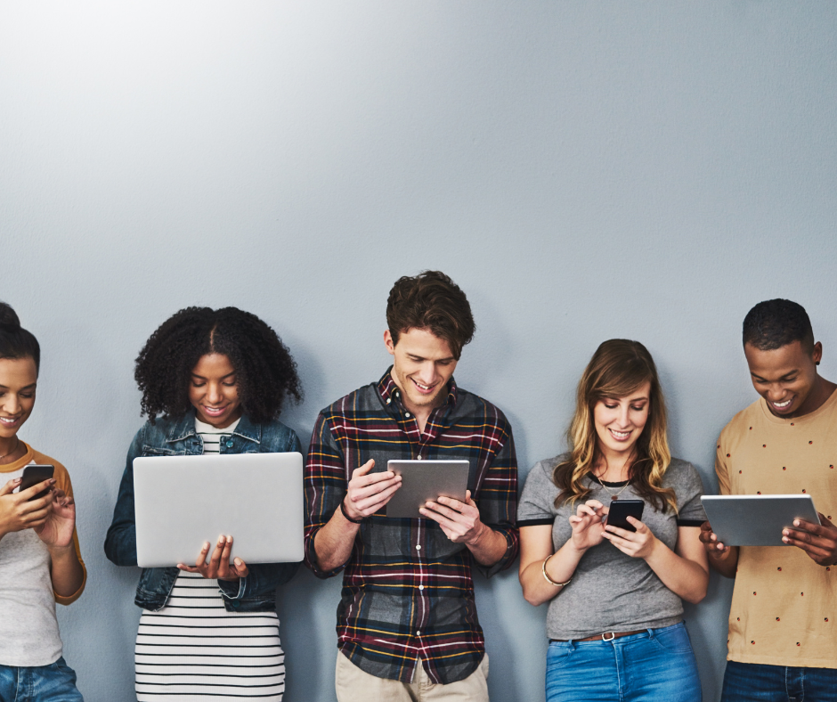 Five young adults of diverse backgrounds stand against a grey wall, smiling while absorbed in a laptop, tablets and smartphones, illustrating how the Tech Scheme puts cutting‑edge devices within reach through employee benefits.